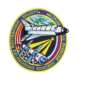 May include: A blue and yellow embroidered patch with the text "Wilcutt - 106 - Altman" and "Mastraccio - Malenchenko - Morukov - Lu Burbank" around the edge. The patch features a space shuttle, a space station, and stars.