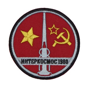 Pode incluir: Um remendo bordado em vermelho, branco e dourado com um foguete, uma foice e um martelo, e duas estrelas. O texto "ИНТЕРКОСМОС 1980" está escrito em preto numa faixa branca em volta do remendo.