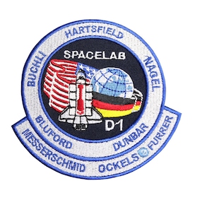 May include: A blue and white embroidered patch with the text "SPACELAB D1" in the centre. The patch also features the names of the astronauts who flew on the mission: "Buchi, Hartsfield, Nagel, Bluford, Messerschmid, Ockels, Dunbar, Furrer".
