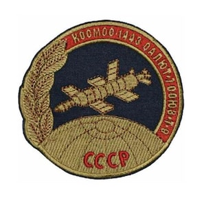 Op de afbeelding: Een ronde, geborduurde patch met een rode rand en gouden letters. De patch toont een gestileerde afbeelding van een ruimtevaartuig dat een planeet omcirkelt, met de tekst "KOOMO 1443 ДАЛЬТ 7 10008-178 CCCP" rond de rand.