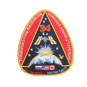 May include: A triangular patch with a red, white and blue design. The patch features a black background with a white starburst, a blue Earth, and a space shuttle. The text "ROMANENKO-FORD MARSHBURN", "CHOVICKIY HADFIELD-TARELKIN", "OFF THE EARTH", and "FOR THE EARTH" is written around the perimeter of the patch. The number "34" is in the centre of the patch.