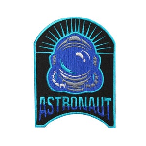 以下が含まれることがあります： 黒の背景に、青と銀の宇宙飛行士のヘルメット。青とターコイズ色の文字で「ASTRONAUT」と書かれている。