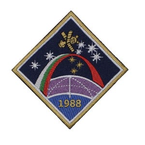 May include: Embroidered patch with a diamond shape, featuring a space-themed design. It includes a satellite, stars, a rainbow arc, and the year 1988. The patch has a navy blue background with gold trim.