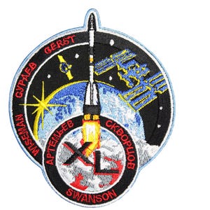 Puede incluir: Un parche circular bordado con fondo negro y un cohete blanco lanzándose al espacio. El parche presenta una Tierra azul y blanca, una estrella amarilla y los nombres de los astronautas: Wiseman, Artemyev, Gerst, Skvortsov, Swanson y Gulaev.