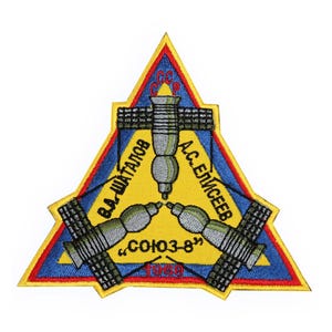 May include: A yellow triangular patch with a blue border and red trim. The patch features three space stations connected in a triangular shape. The text "CCCP" is at the top of the triangle. The text "Г.А. ШАТАЛОВ" is on the left side of the triangle. The text "А.С. ЕЛИСЕЕВ" is on the right side of the triangle. The text "СОЮЗ-8" is at the bottom of the triangle.