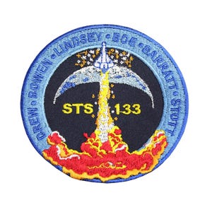 May include: Embroidered patch depicting a rocket launch, with the text "STS .133". The patch has a blue border with crew names. The rocket's flames are red and yellow, set against a dark blue background.