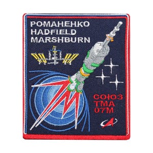 May include: Embroidered patch with a space shuttle launch design, featuring the names ROMAHENKO, HADFIELD, and MARSHBURN. The patch includes the text CO103 TMA 07M, a space station, and a stylized rocket against a navy blue background with red and white accents.