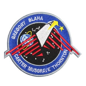 May include: A blue and white embroidered patch with the text "Gregory Blaha Carter Musgrave Thornton" around the edge. The patch features a stylized image of a rocket ship and a planet.