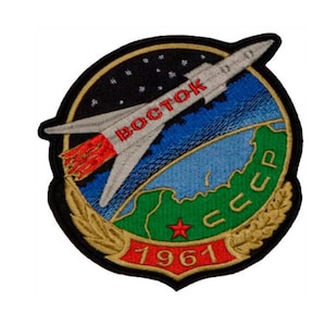May include: A black and gold embroidered patch featuring a spacecraft with the text "BOSTOK" and "CCCP" in red, flying over a green and blue globe with a red star. The year "1961" is written in red below the globe.