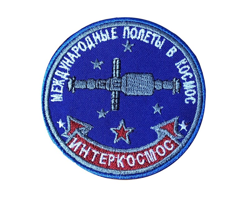 Pode incluir: Um remendo bordado azul com o texto "Международные полеты в космос Интеркосмос" em letras brancas e vermelhas. O remendo apresenta uma imagem estilizada de uma esta&ccedil;&atilde;o espacial com estrelas no fundo.