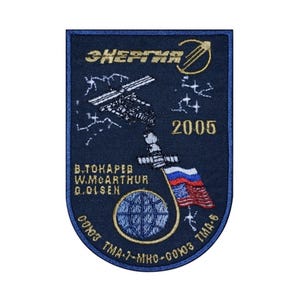 May include: A blue embroidered patch with the text "ЭНЕРГИЯ" and "2005" in gold lettering. The patch features a depiction of the International Space Station, a Russian flag, and an American flag. The names "В.ТОКАРЕВ", "W.MCARTHUR", and "G.OLSEN" are also included in gold lettering.