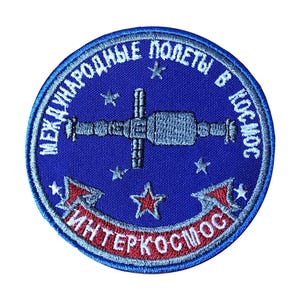 May include: A blue embroidered patch with the text "Международные полеты в космос Интеркосмос" in white and red lettering. The patch features a stylized image of a space station with stars in the background.