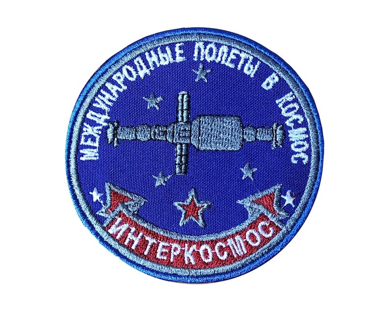 Pode incluir: Um remendo bordado azul com o texto "Международные полеты в космос Интеркосмос" em letras brancas e vermelhas. O remendo apresenta uma imagem estilizada de uma esta&ccedil;&atilde;o espacial com estrelas no fundo.