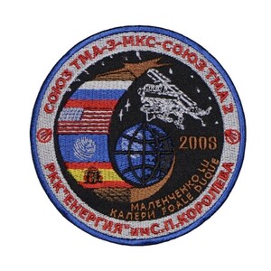 May include: A circular embroidered patch with a blue border and red text. The text reads "Soyuz TMA-3-MKS-Soyuz TMA 2" and "RKK Energia IMS.L.Koroleva". The center of the patch features a black background with a white space shuttle, a globe, and three flags: the American flag, the Russian flag, and the flag of the European Space Agency. The year "2003" is printed in white on the black background. The text "Malenchenko Lu Kaleri Foale Duque" is printed in red on the blue border.