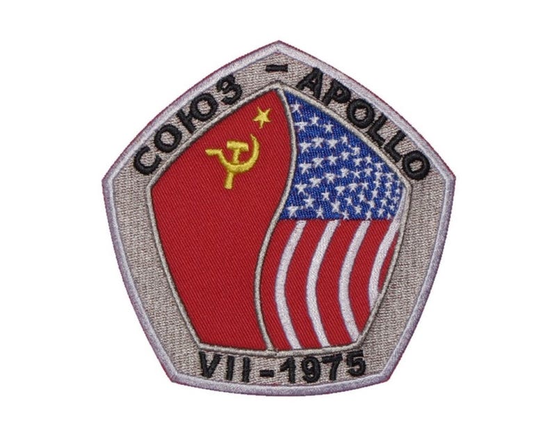 Peut inclure: Un &eacute;cusson brod&eacute; argent&eacute; et gris avec le texte "Soyouz - Apollo VII-1975" et un drapeau rouge et blanc avec une faucille et un marteau et des &eacute;toiles.