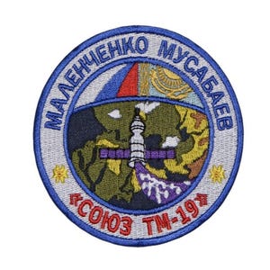 May include: A blue and white embroidered patch with the text "МАЛЕНЧЕНКО МУСАБАЕВ "СОЮЗ ТМ-19" in Cyrillic script. The patch features a stylised image of a space station and a globe.