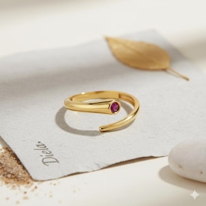 Può includere: Un anello color oro con una piccola gemma rossa rotonda incastonata al centro. L'anello ha un design aperto e unico. L'anello è su un pezzo di carta con la scritta "Diela" stampata.