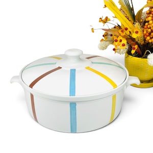 May include: A white ceramic casserole dish with a matching lid, featuring two side handles and a central knob. The exterior and lid are decorated with vertical painted stripes in shades of light blue, yellow, and brown, evenly spaced around the body.
