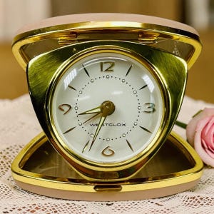 May include: A Westclox travel alarm clock with a gold-tone metal frame and a light pink exterior case. The round white clock face features gold-tone numerals at the 12, 3, 6, and 9 positions, thin gold-tone hands, and a clear protective cover.