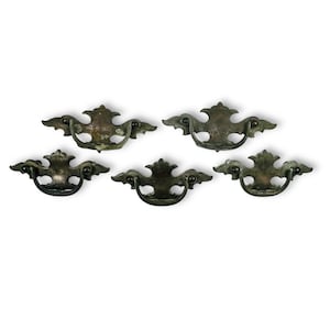 May include: Five antique bronze drawer pulls with a decorative, ornate design. Each pull features a curved handle and a detailed, symmetrical shape with wing-like extensions. The pulls are arranged against a white background.