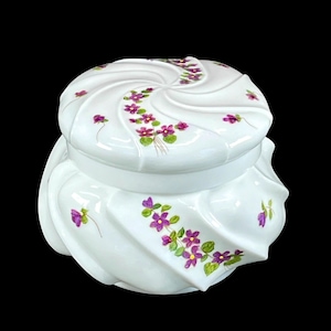 May include: A white ceramic jar with a lid, featuring a spiral design and adorned with hand-painted purple flowers and green leaves. The jar has a round shape and is set against a black background.