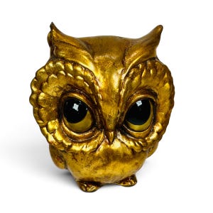May include: A gold-colored owl figurine with a textured, metallic finish. The owl features large, prominent black eyes with reflective highlights, a small beak, and detailed feather patterns around the face and body.