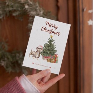 May include: A Christmas card featuring a dachshund wearing a Santa hat standing next to a decorated Christmas tree with presents underneath. The card reads "Merry Christmas and Happy New Year".