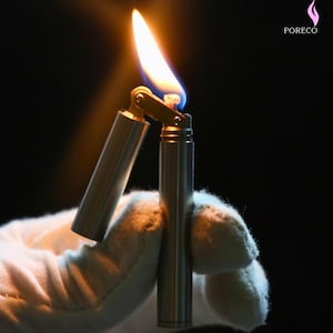 K&ouml;nnte beinhalten: Ein silberfarbenes, zylindrisches Feuerzeug mit einer Flamme. Das Feuerzeug ist ge&ouml;ffnet, wobei der obere Teil vom Hauptk&ouml;rper abgewinkelt ist. Die Flamme ist gelb und orange mit einem blauen Sockel. Das Feuerzeug wird von einer behandschuhten Hand vor dunklem Hintergrund gehalten.