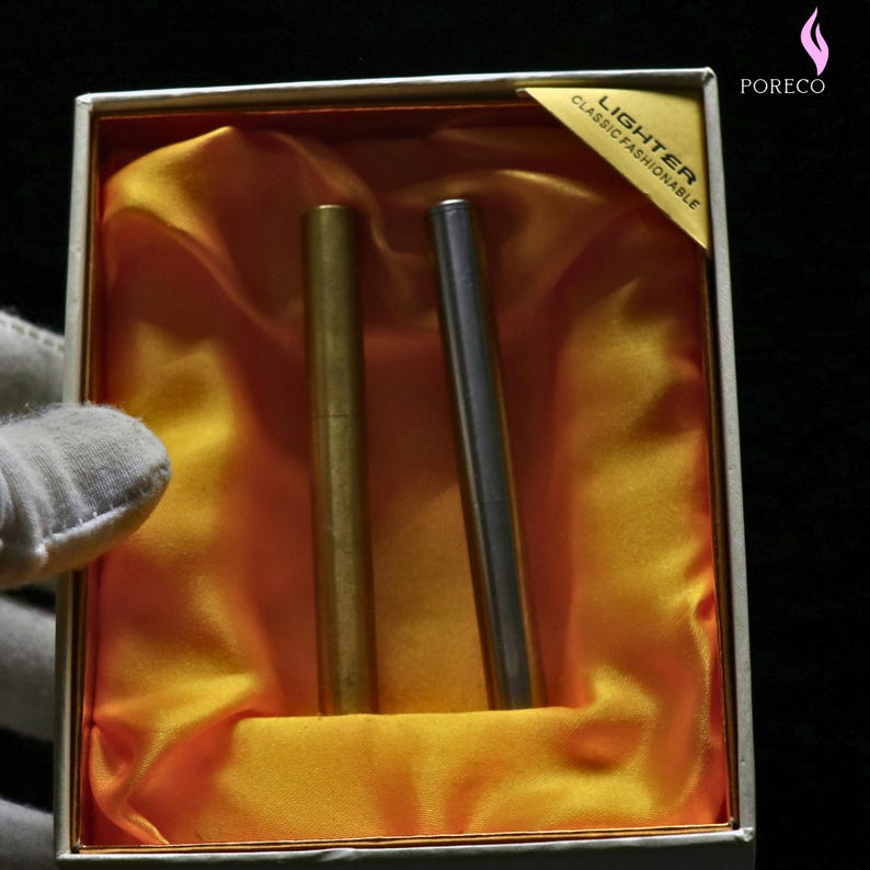 K&ouml;nnte beinhalten: Eine Geschenkbox mit zwei schlanken, zylindrischen Feuerzeugen. Eines ist goldfarben, das andere silberfarben. Sie liegen auf orangefarbenem Satin in einer wei&szlig;en Box. Auf der Box befindet sich ein goldenes Etikett mit der Aufschrift "LIGHTER CLASSIC FASHIONABLE".
