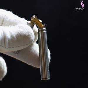 K&ouml;nnte beinhalten: Ein silberfarbenes, zylindrisches Feuerzeug mit einem messingfarbenen Scharnier und Oberteil. Es wird in einem wei&szlig;en Handschuh vor schwarzem Hintergrund gehalten. Der Markenname "PORECO" ist sichtbar. Das Design deutet auf Funktionalit&auml;t und Stil hin.
