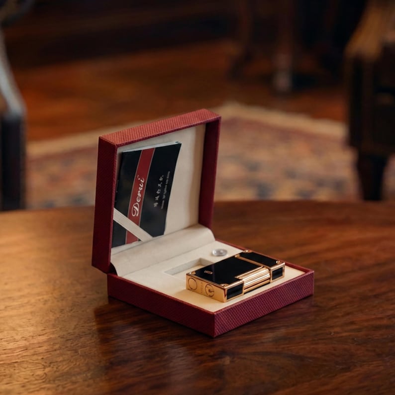 May include: A black and gold lighter in a red presentation box with a cream interior. The box contains a card with the brand name "Derui". The lighter is on a wooden surface.