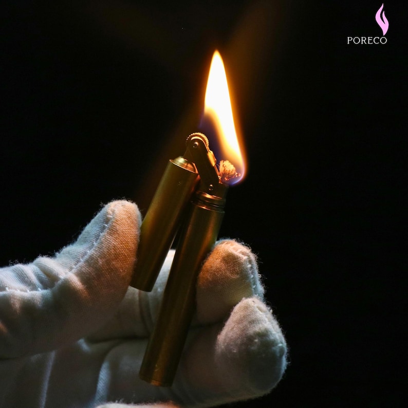 K&ouml;nnte beinhalten: Ein brennendes, im Vintage-Stil gehaltenes Messingfeuerzeug mit langer, heller Flamme. Das Feuerzeug wird in einer behandschuhten Hand vor schwarzem Hintergrund gehalten. Der Markenname "PORECO" ist oben rechts sichtbar.