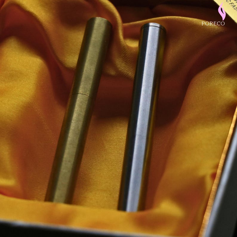 K&ouml;nnte beinhalten: Zwei metallene stiftf&ouml;rmige Objekte, eines goldfarben und eines silberfarben, werden in einer Schachtel mit gelbem Satinfutter pr&auml;sentiert. Die Stifte haben ein schlankes, zylindrisches Design. Der goldene Stift hat eine sichtbare Naht.