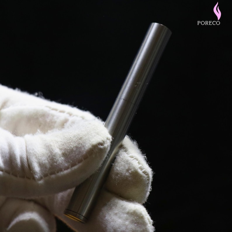K&ouml;nnte beinhalten: Ein silberfarbenes, zylindrisches Metallobjekt, m&ouml;glicherweise ein Stift, wird von einer behandschuhten Hand vor einem schwarzen Hintergrund gehalten. Das Objekt hat eine glatte, reflektierende Oberfl&auml;che. Der Markenname "PORECO" ist in der oberen rechten Ecke sichtbar.