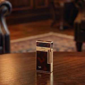May include: A rectangular lighter with a gold-coloured frame and a tortoiseshell pattern on the front and top. The lighter has the word "Derai" and "Lighter" printed on it. The bottom of the lighter has a "D" logo.