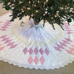 May include: A white Christmas tree skirt with a quilted design featuring pink diamond shapes. The skirt has a scalloped lace edge and is placed under a decorated Christmas tree with ornaments.