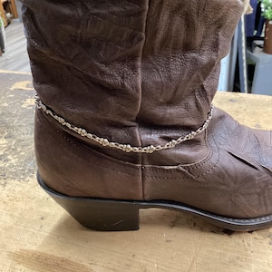 May include: A brown leather boot with a silver chain ankle bracelet. The boot has a worn, textured appearance and a low heel. The silver chain is adorned with small, decorative elements.