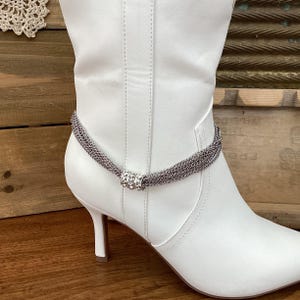 May include: A silver-toned ankle bracelet with multiple strands of chain and a decorative center piece with clear rhinestones. The bracelet is shown on a white leather boot with a stiletto heel.