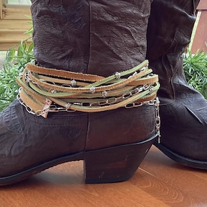 May include: A pair of brown leather cowboy boots with a pointed toe and stacked heel. The boots are adorned with a decorative ankle wrap featuring leather straps, silver chain, and small metal accents. The boots are displayed on a wooden surface.