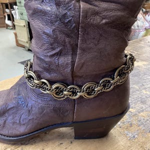 May include: A brown leather boot featuring a textured, wrinkled surface and a low heel. Wrapped around the ankle area is a decorative accessory consisting of multiple layers of interconnected brass-toned metal chains and circular links.
