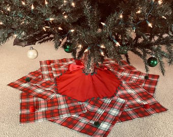 May include: A Christmas tree skirt with a red and white plaid pattern, accented with green and red pom-poms. The skirt has a red center with a green trim and is placed under a decorated Christmas tree with ornaments and lights.