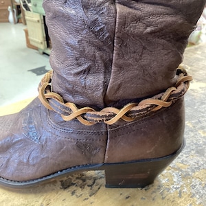 May include: Close-up of a brown leather cowboy boot with a decorative braided leather ankle wrap. The boot has a worn, textured appearance with embossed floral patterns. The heel is approximately 1.5 inches high.