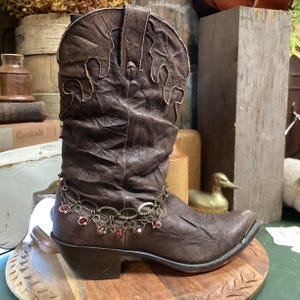 May include: A brown leather cowboy boot with a pointed toe and decorative stitching. The boot features a chain with red embellishments around the ankle. The boot is on a wooden surface.