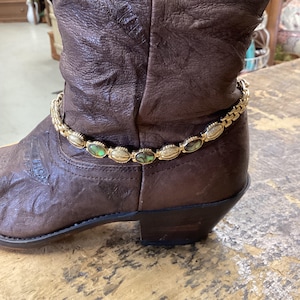 May include: A dark brown leather cowboy boot adorned with a gold-toned chain ankle bracelet. The bracelet features alternating oval links, with some links containing iridescent green and brown shell inlays and others showing a textured gold finish.
