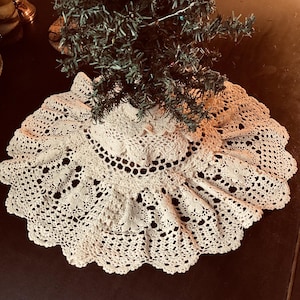 May include: A circular, off-white crocheted tree skirt featuring an intricate floral pattern with scalloped edges. The fabric is crafted with detailed needlework, creating a delicate, lacy texture that spreads outward from the center base.