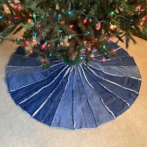 May include: A circular Christmas tree skirt constructed from multiple vertical panels of blue denim fabric. The panels feature varying shades of blue, exposed frayed seams, and visible pocket details, arranged in a radial pattern around the base.