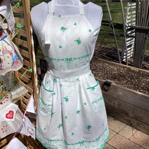 May include: White apron with a floral and bow pattern in turquoise. The apron has two pockets, a waist tie, and a lace trim. The apron is displayed on a mannequin in an outdoor setting.