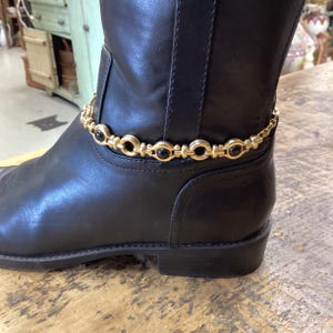 May include: A black leather boot adorned with a gold-toned metal chain ankle bracelet. The bracelet features a repeating pattern of circular gold links alternating with smooth, round black stones, encircling the lower part of the boot shaft.