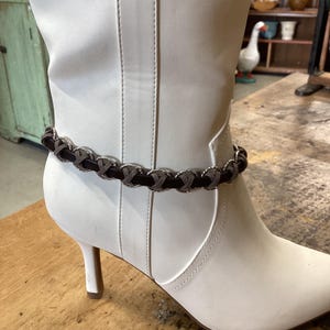May include: A white leather ankle boot with a pointed toe and a small heel. A decorative ankle bracelet with a dark brown band and silver-colored accents encircles the boot.