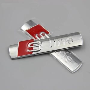Op de afbeelding: Twee zilveren auto-emblemen met de tekst "S line" in reliëf. Het bovenste embleem heeft een rode achtergrond met een gestileerde "S" in het midden.